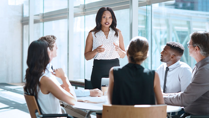 business team working at a conference table. An african american woman is standing leading the conversation.