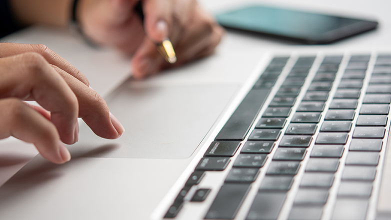 A close up of a person's hand using the trackpad on a laptop. The hand is in focus while the laptop is slightly out of focus. A smartphone is in the background.