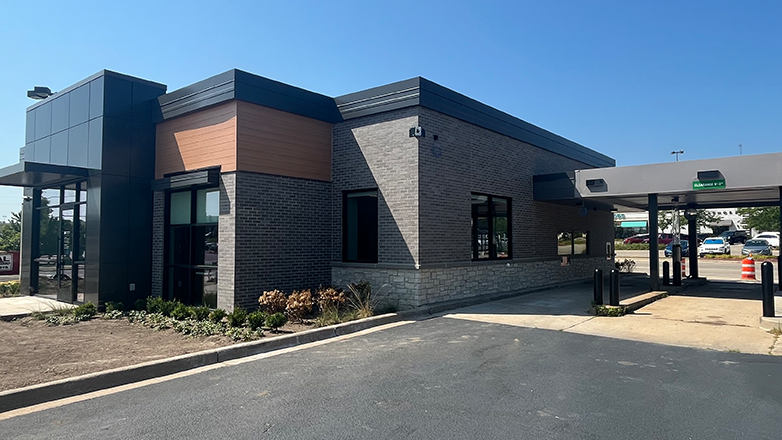 A modern brick building with a drive-thru bank. The building has large windows and a black awning over the entrance. There is a drive-thru lane on the right side of the building. There are cars parked in the parking lot.