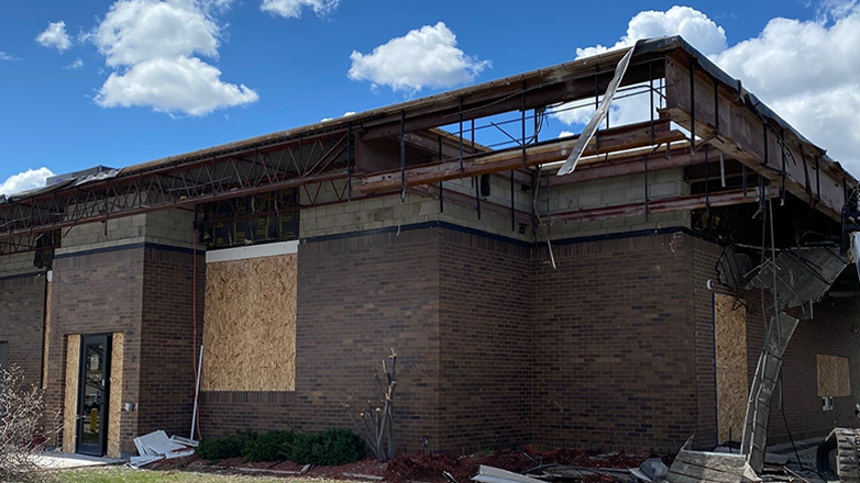 A brick building with a partially collapsed roof. The roof is supported by scaffolding and the building has boarded up windows. There is a blue sky with white clouds in the background..