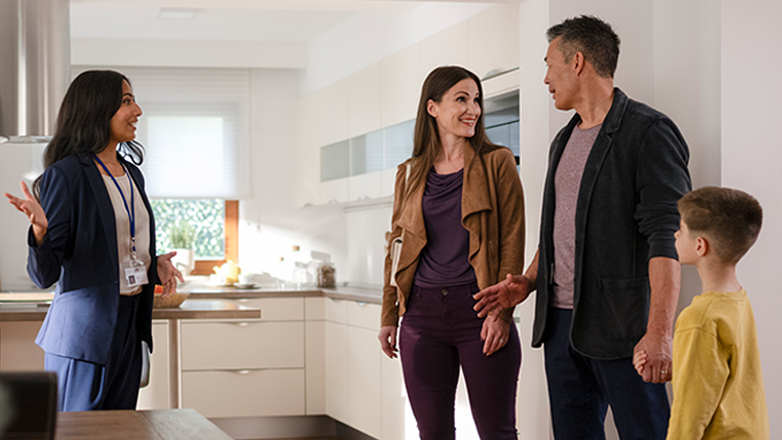 A real estate agent is showing a couple and their child a house. The agent is standing in the kitchen and is talking to the couple. The couple is standing in the living room and is looking at the agent. The child is standing next to the couple and is looking at the house.