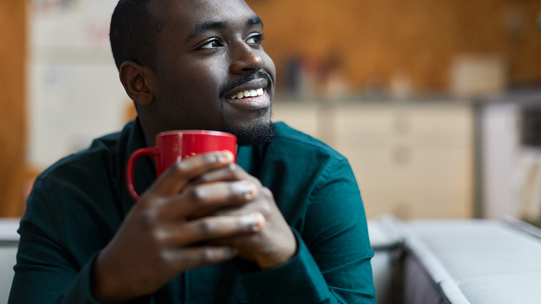 A young African-American man is sitting on a couch and holding a red coffee mug. He is looking off to the side and smiling.