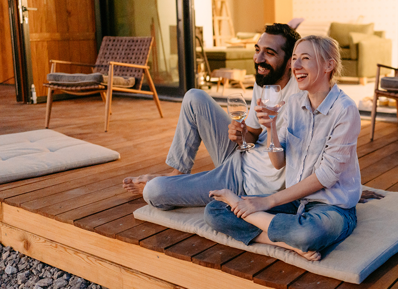 A man and woman sit facing forward with big smiles on a wooden deck, each holding a glass of white wine. They are angled towards each other, and the woman's bare foot is touching the man's calf. The evening sun is casting a warm glow on them.