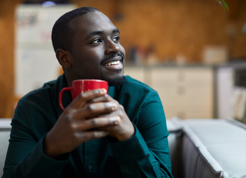 A young African-American man is sitting on a couch and holding a red coffee mug. He is looking off to the side and smiling.