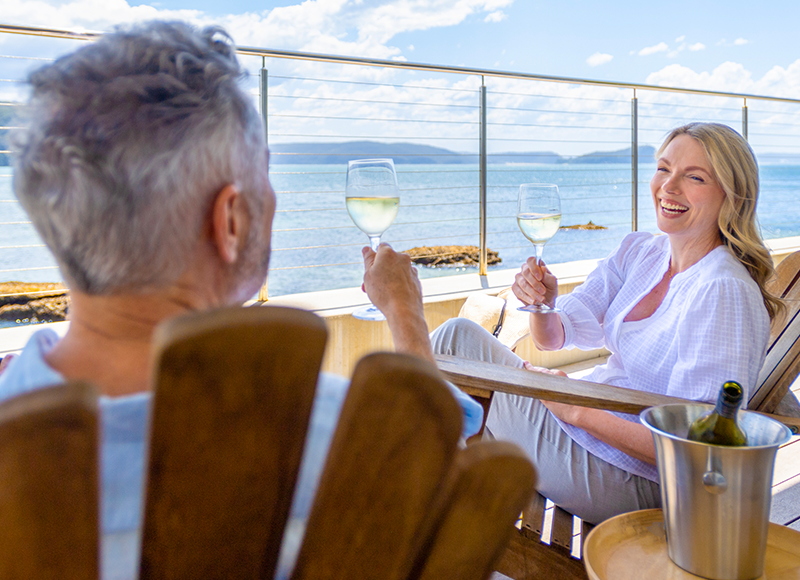 A man and woman are sitting on a balcony overlooking the ocean, both raising a glass of white wine to each other. The woman is laughing while the man is looking away from the camera. A bottle of wine sits in a bucket on a small table beside them.