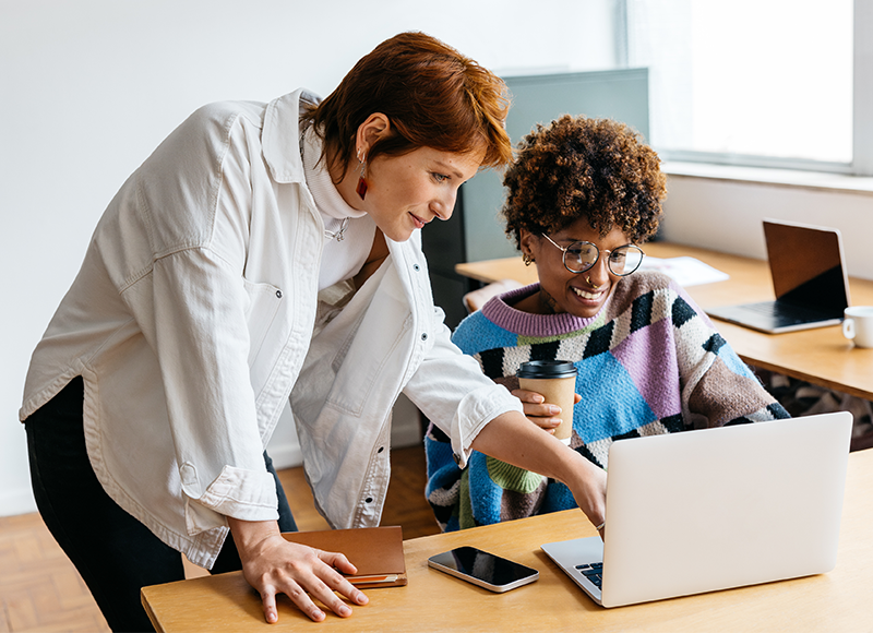 Two women are working together in an office. The woman in the white shirt is leaning over the shoulder of the woman sitting at the desk, pointing at the screen of her laptop. The woman sitting is holding a cup of coffee.