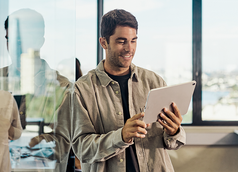 A man stands in an office, looking at a tablet in his hands. He is smiling, and the office has large windows looking out to the city. The image suggests a positive and professional work environment.