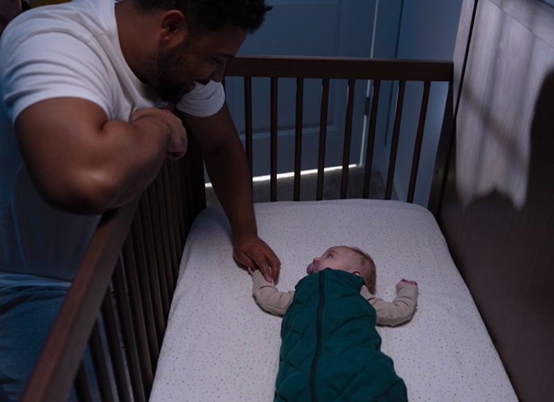 A father beams at his little one, nestled in the crib, snug in a sleep sack. He leans in, gently grasping the tiny hand, a moment of pure connection.