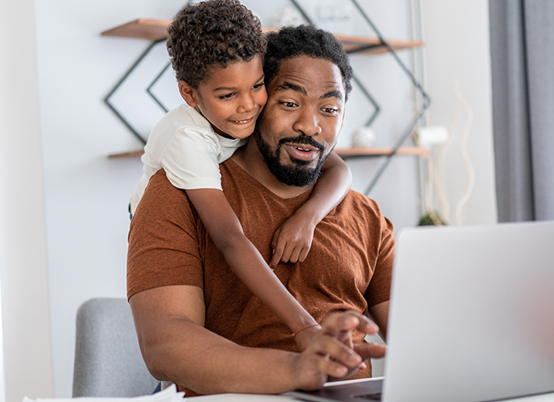 An african american father and son are looking at a laptop together. The father is smiling and pointing at something on the screen. The son is smiling and hugging his father.