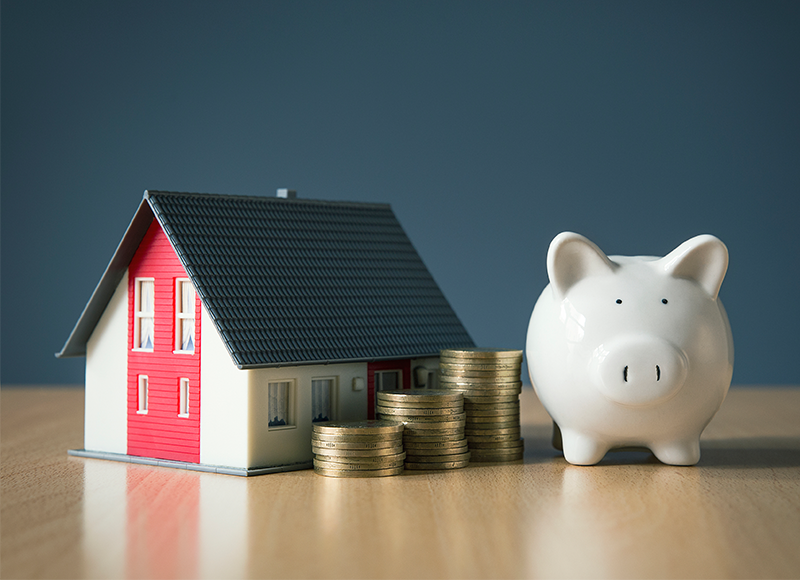 A piggy bank sits next to a small model house. Stacks of coins are in front of the house.