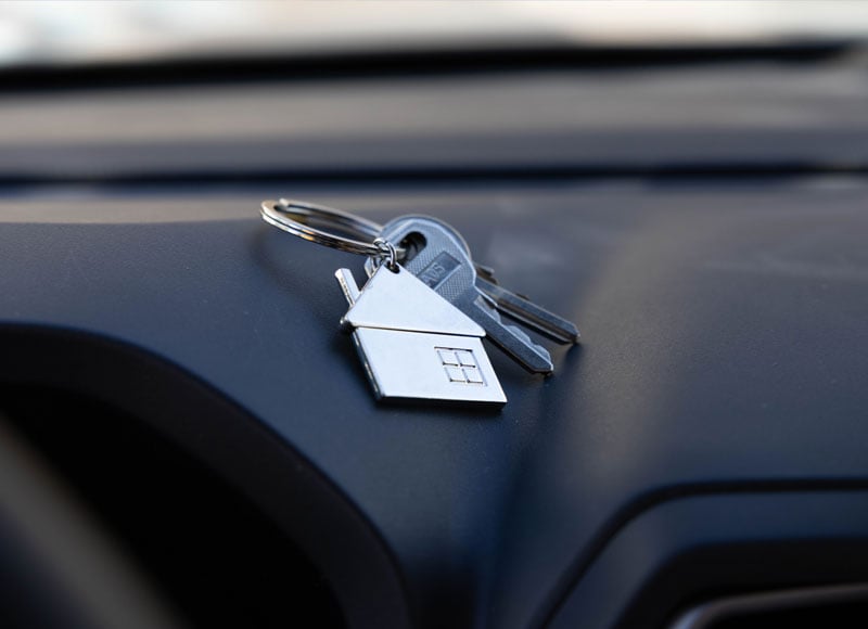 A close-up of a silver metal keychain in the shape of a house, with two house keys attached. The keychain rests on the dark dashboard of a car.