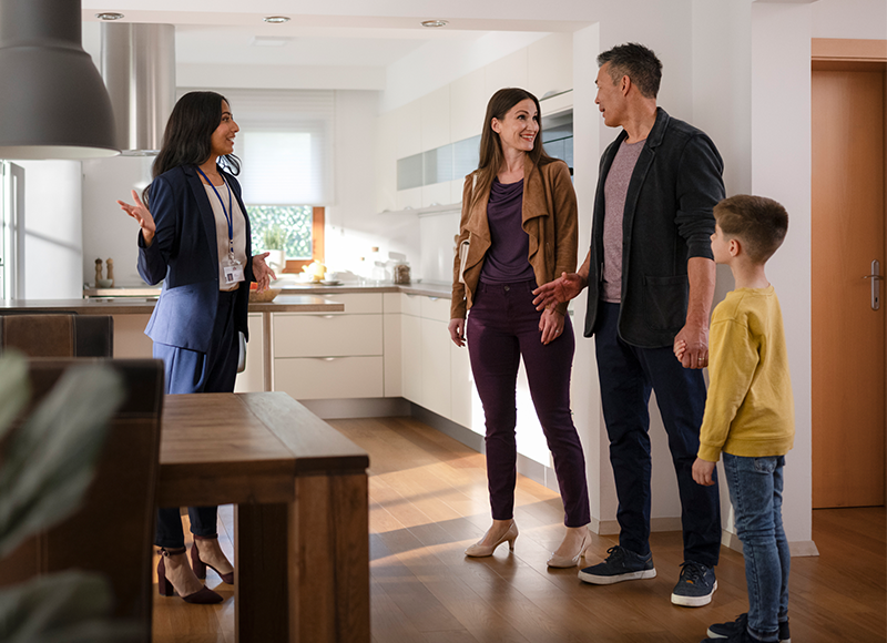 A real estate agent is showing a couple and their child a house. The agent is standing in the kitchen and is talking to the couple. The couple is standing in the living room and is looking at the agent. The child is standing next to the couple and is looking at the house.