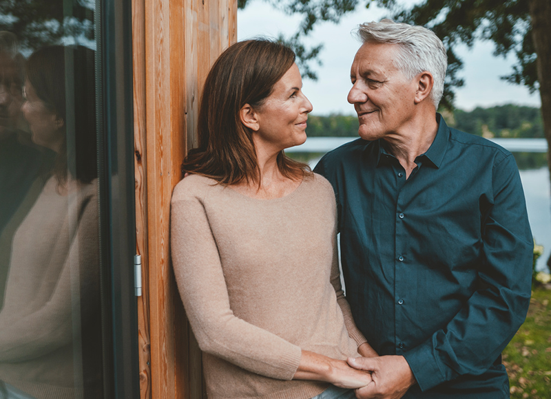 A mature man and woman, embracing gently, gaze into each other's eyes. They are standing near a wooden structure by a lake.